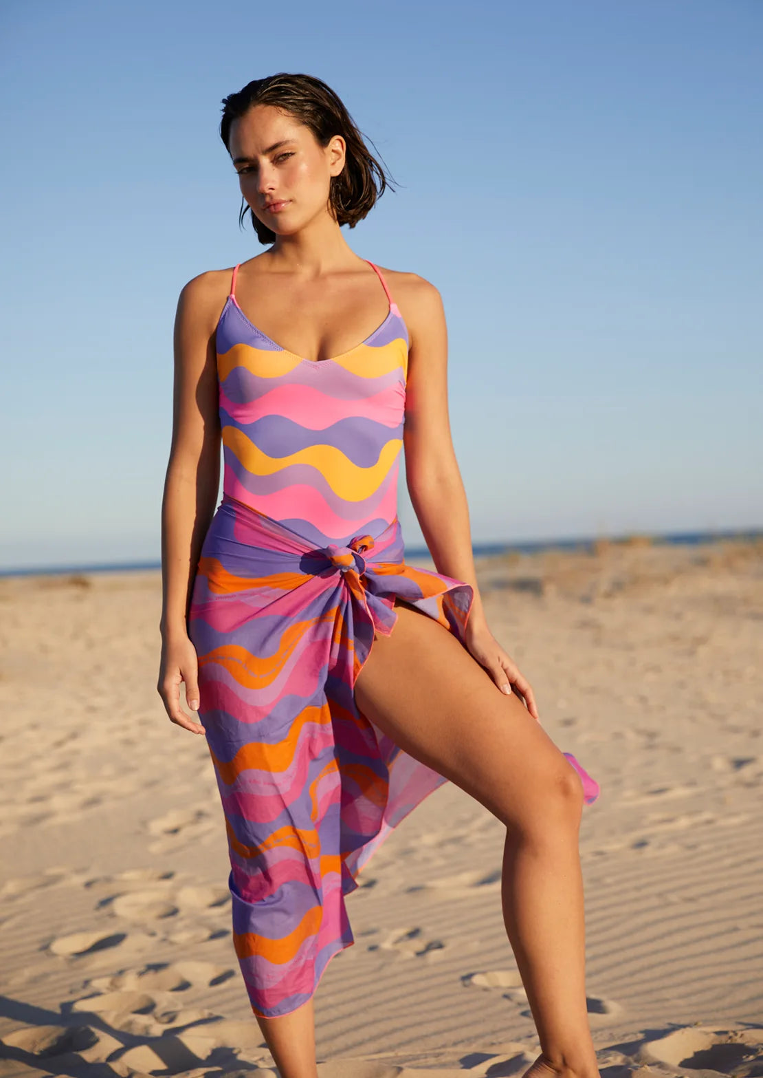 Bañador estampado de olas en color lila, fucsia, morado y naranja. Tirantes espagueti cruzados en la espalda. Bañador Waves de To the Moon para mujer y teen con Pareo a juego.